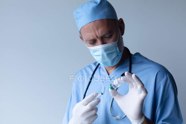 Médecin masculin caucasien portant un masque facial debout et une seringue de remplissage. professionnel de la santé hygiène du personnel de santé pendant le coronavirus covid 19 pandémie. — Photo de stock