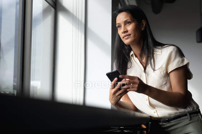 Femme d'affaires mixte debout par la fenêtre à l'aide d'un smartphone dans un bureau moderne. entreprise moderne bureau lieu de travail technologie. — Photo de stock