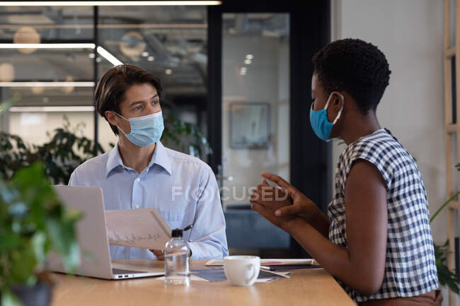 Diverse business people wearing face masks sitting using laptop going through paperwork in office. hygiene in business office workplace during covid 19 coronavirus pandemic. — Stock Photo