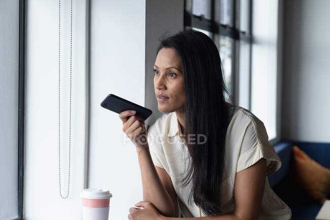 Mulher de negócios de raça mista em pé pela janela usando smartphone no escritório moderno. negócio moderno escritório tecnologia do local de trabalho. — Fotografia de Stock