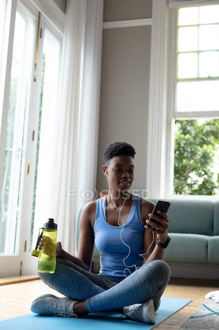 Donna afroamericana in possesso di bottiglia d'acqua utilizzando smartphone a casa. stare a casa in isolamento personale in quarantena — Foto stock