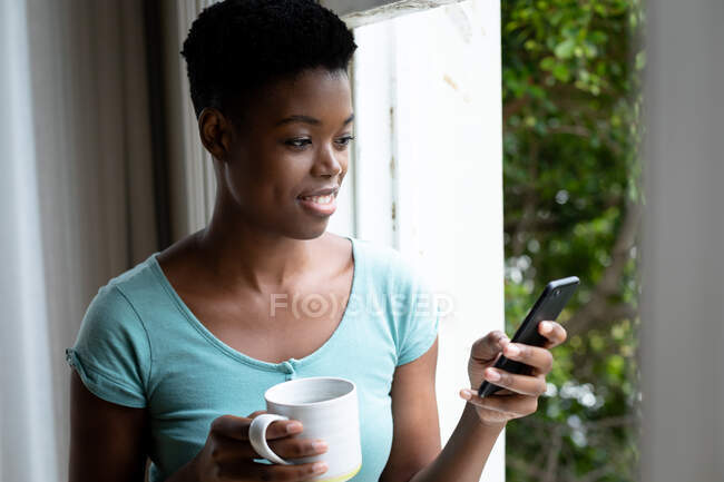 Porträt einer Afroamerikanerin, die zu Hause eine Kaffeetasse mit dem Smartphone in der Hand hält. Zu Hause bleiben in Selbstisolierung in Quarantäne — Stockfoto