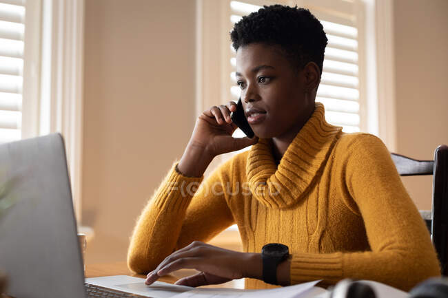 African american woman using laptop talking on smartphone in kitchen. staying at home in self isolation during quarantine lockdown. — Stock Photo