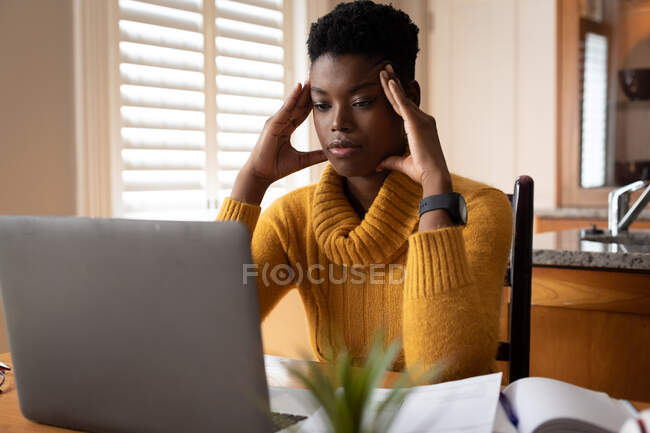 Afro-americana usando laptop esfregando seus templos na cozinha. ficar em casa em auto-isolamento durante o confinamento de quarentena. — Fotografia de Stock