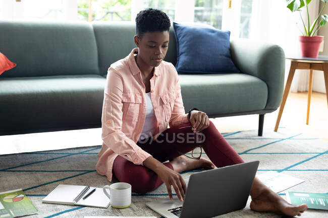 African american woman sitting on floor using laptop drinking coffee working from home. staying at home in self isolation during quarantine lockdown. — Stock Photo