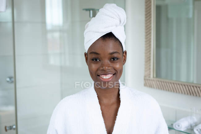 Retrato de mujer afroamericana en albornoz sonriendo en el baño en casa. permaneciendo en casa en aislamiento en cuarentena - foto de stock
