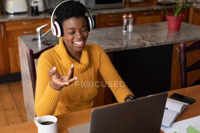 Mujer afroamericana usando auriculares haciendo videollamadas usando laptop en la cocina. permanecer en casa en aislamiento durante el bloqueo de cuarentena. - foto de stock