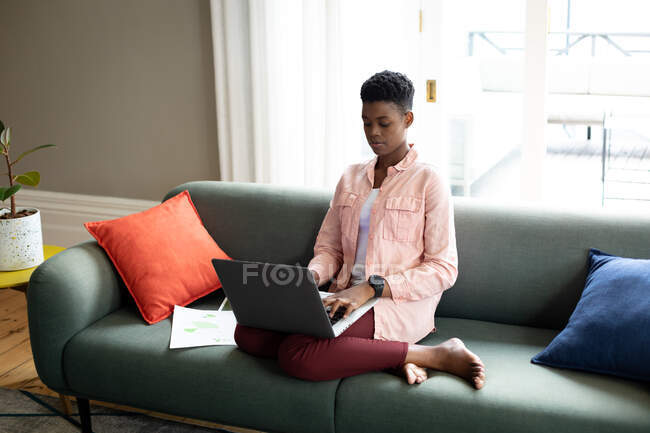 Mujer afroamericana sentada en el sofá usando el portátil hablando en el teléfono inteligente que trabaja desde casa. permanecer en casa en aislamiento durante el bloqueo de cuarentena. - foto de stock