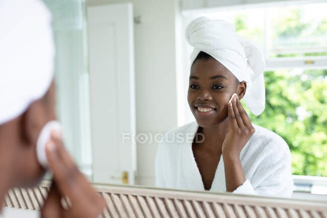 Mujer afroamericana en albornoz quitando el maquillaje mientras se mira en el espejo en el baño. permaneciendo en casa en aislamiento en cuarentena - foto de stock