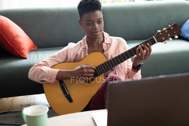 Mulher afro-americana tocando guitarra enquanto olha para o laptop em casa. ficar em casa em auto-isolamento em quarentena — Fotografia de Stock