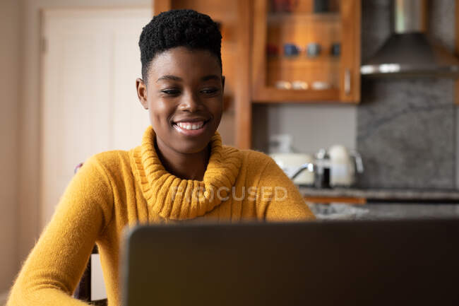 Mujer afroamericana usando laptop y sonriendo en la cocina. permanecer en casa en aislamiento durante el bloqueo de cuarentena. - foto de stock