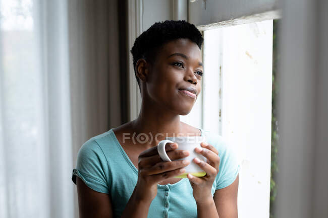 Portrait de femme afro-américaine tenant une tasse de café regardant par la fenêtre à la maison. rester à la maison en isolement personnel en quarantaine — Photo de stock