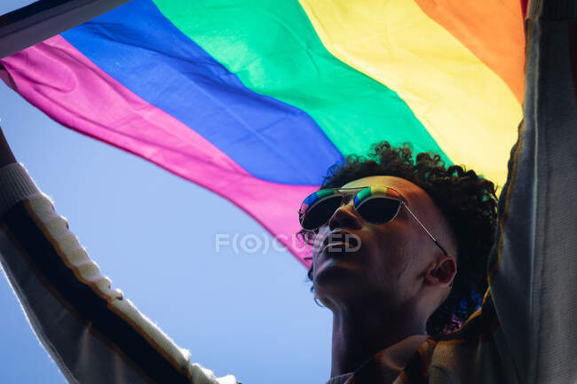 Homme de race mixte debout sur le toit tenant le drapeau arc-en-ciel. genre fluide identité lgbt concept d'égalité raciale. — Photo de stock