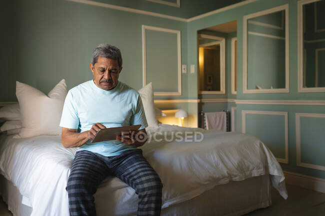 Старший африканский американец, сидящий на кровати с цифровым планшетом в спальне. Пенсионный образ жизни в самоизоляции во время пандемии коронавируса. — стоковое фото