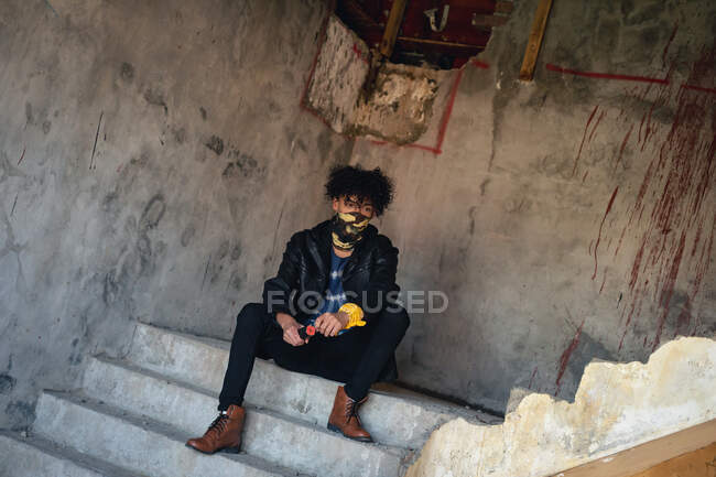 Uomo di razza mista in un edificio vuoto che indossa una maschera facciale seduta sulle scale. genere fluido lgbt identità concetto di uguaglianza razziale. — Foto stock