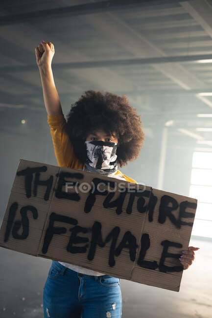 Mujer de raza mixta con máscara facial sosteniendo un cartel de protesta levantando la mano. género fluido lgbt identidad concepto de igualdad racial. - foto de stock