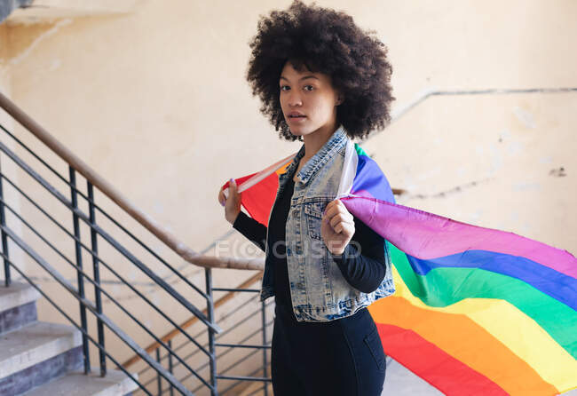 Mujer de raza mixta sosteniendo una bandera del arco iris mirando a la cámara. género fluido lgbt identidad concepto de igualdad racial. - foto de stock