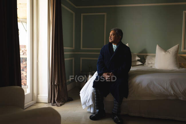 Hombre afroamericano mayor sentado en una cama en un dormitorio. estilo de vida de jubilación en aislamiento durante el coronavirus covid 19 pandemia. - foto de stock