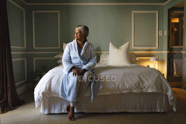 Mulher afro-americana sênior sentada em uma cama em um quarto de dormir. estilo de vida de aposentadoria em auto-isolamento durante coronavírus covid 19 pandemia. — Fotografia de Stock