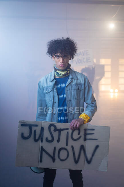 Mixed race man wearing face mask holding protest sign raising fist. with protesters in the background. gender fluid lgbt identity racial equality concept. — Stock Photo