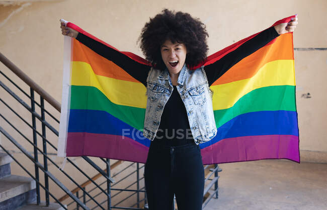 Mujer de raza mixta sosteniendo la bandera del arco iris gritando a la cámara. género fluido lgbt identidad concepto de igualdad racial. - foto de stock