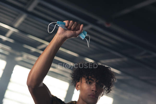 Mixed race man in an empty building raising a fist with face mask in it. gender fluid lgbt identity racial equality concept. — Stock Photo