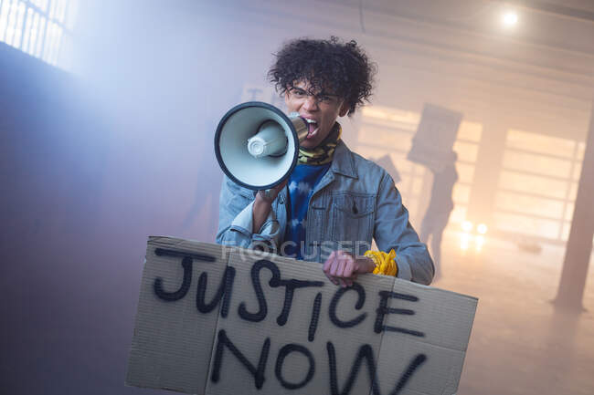 Hombre de raza mixta gritando en megáfono sosteniendo un cartel de protesta. con manifestantes en el fondo. género fluido lgbt identidad concepto de igualdad racial. - foto de stock