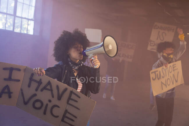 Mujer de raza mixta gritando en megáfono sosteniendo un cartel de protesta. con manifestantes en el fondo. género fluido lgbt identidad concepto de igualdad racial. - foto de stock