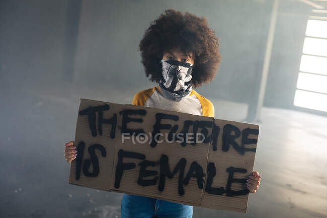 Mulher de raça mista usando máscara facial segurando sinal de protesto. gênero fluido lgbt identidade conceito de igualdade racial. — Fotografia de Stock