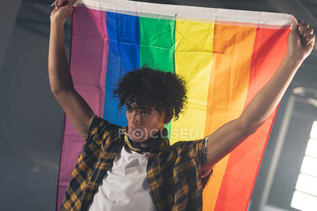 Hombre de raza mixta parado en la azotea sosteniendo la bandera del arco iris. género fluido lgbt identidad concepto de igualdad racial. - foto de stock