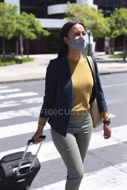 African american woman wearing face mask with trolley bag crossing the street. lifestyle living during coronavirus covid 19 pandemic. — Stock Photo