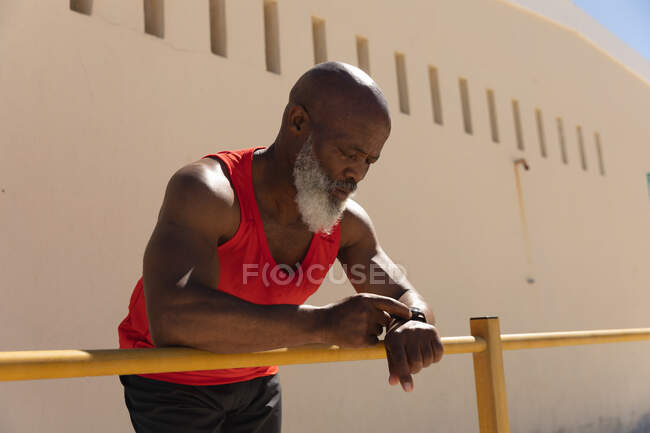 S'adapter homme afro-américain senior exercice en utilisant smartwatch appuyé sur la clôture au soleil. saine retraite technologie communication extérieur forme physique mode de vie. — Photo de stock