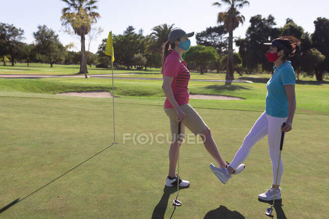 Two caucasian women wearing face masks greeting on golf course by touching ankles. sport leisure hobbies golf healthy outdoor lifestyle hygiene during coronavirus covid 19 pandemic. — Stock Photo
