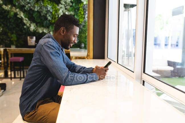 Hombre afroamericano sentado en un café usando un teléfono inteligente y sonriendo. hombre de negocios en la salida en la ciudad. - foto de stock