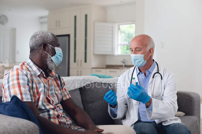 Senior caucasian male doctor preparing vaccine for male patient both wearing face masks at home. healthcare hygiene protection during coronavirus covid 19 pandemic. — Stock Photo
