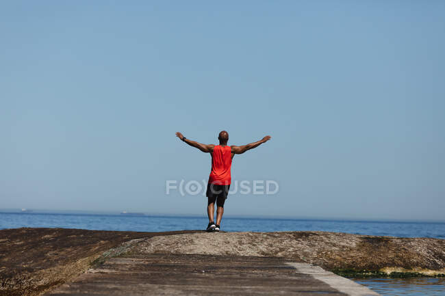 Rückansicht eines fitten älteren afrikanisch-amerikanischen Mannes, der mit ausgestreckten Armen auf dem Meer trainiert. gesunder Lebensstil im Freien im Ruhestand. — Stockfoto