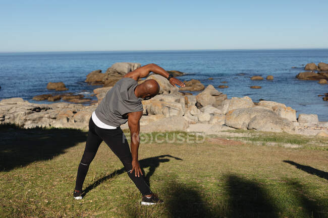 Visão traseira do homem americano africano sênior apto que exercita-se ao ar livre pelo mar que estica. aposentadoria saudável ao ar livre fitness lifestyle. — Fotografia de Stock