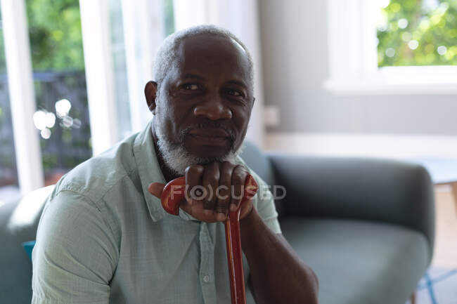 Портрет старшого афроамериканського чоловіка, який сидить у вітальні, спираючись на ходячу палицю. Перебуваючи вдома в ізоляції під час карантину.. — стокове фото