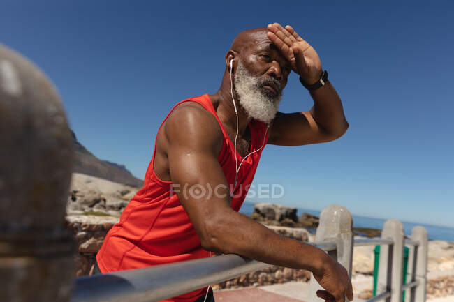 Ajuste o homem americano africano sênior que usa fones de ouvido no sol pelo mar que esfrega a testa. tecnologia de aposentadoria saudável comunicação ao ar livre fitness lifestyle. — Fotografia de Stock