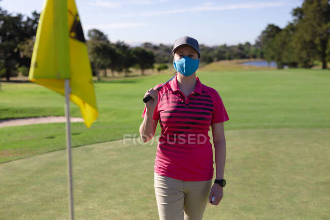 Retrato de mujer caucásica con máscara facial que sostiene el club en el campo de golf. deporte ocio aficiones golf saludable estilo de vida al aire libre higiene durante coronavirus covid 19 pandemia. - foto de stock