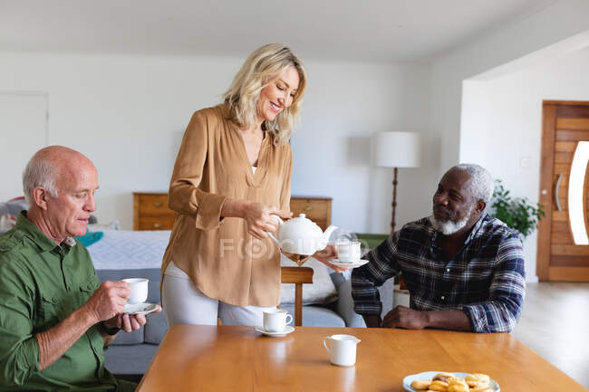 Personas mayores caucásicas y afroamericanas sentadas a la mesa tomando té en casa. senior retiro estilo de vida amigos socializando. - foto de stock