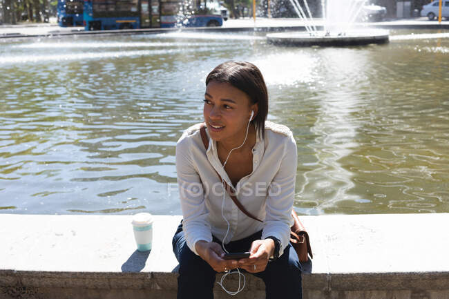 African american woman wearing earphones listening to music while sitting at corporate park. lifestyle living concept during coronavirus covid 19 pandemic. — Stock Photo