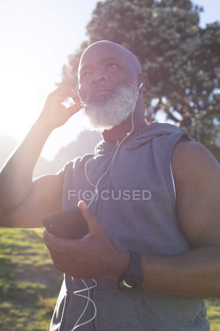 Ajuste homem americano africano sênior segurando smartphone colocando fones de ouvido. tecnologia de aposentadoria saudável comunicação ao ar livre fitness lifestyle. — Fotografia de Stock