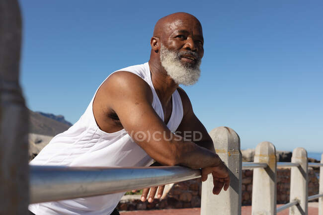 Fit Senior afrikanisch-amerikanischer Mann lehnt am Zaun vor blauem Himmel. gesunde Ruhestand Technologie Kommunikation Outdoor Fitness Lebensstil. — Stockfoto