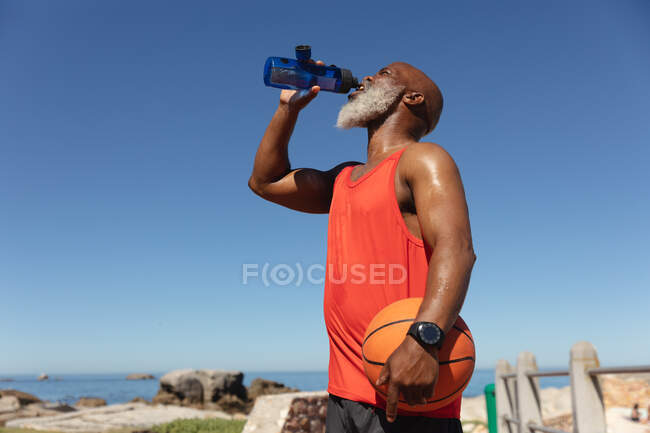 Подходит старшему африканскому американцу по морю, пьющему из бутылки с водой, держащему баскетбол. здоровый образ жизни на свежем воздухе. — стоковое фото