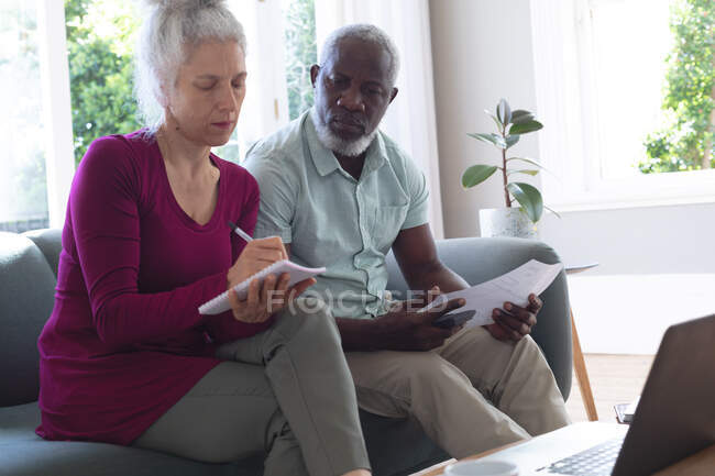 Senior mixed race couple using laptop paying bills together in living room. staying at home in self isolation during quarantine lockdown. — Stock Photo