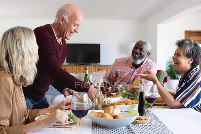 Parejas mayores caucásicas y afroamericanas sentadas a la mesa cenando en casa. senior retiro estilo de vida amigos socializar. - foto de stock