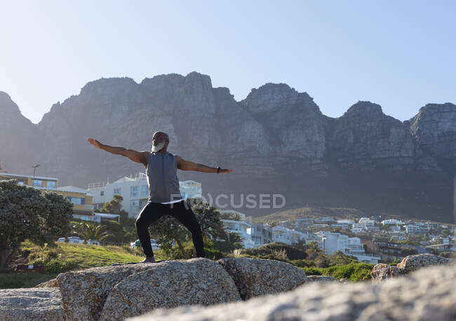 Fit senior african american man standing on rocky coast doing yoga. healthy retirement outdoor fitness lifestyle. — Stock Photo