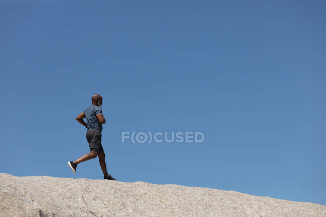 Ajustez l'homme afro-américain senior s'exerçant à courir sur le rocher contre le ciel bleu. retraite saine mode de vie en plein air fitness. — Photo de stock