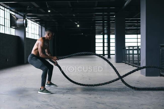Homme afro-américain portant des vêtements de sport luttant contre les cordes dans un bâtiment urbain vide. forme physique urbaine mode de vie sain. — Photo de stock